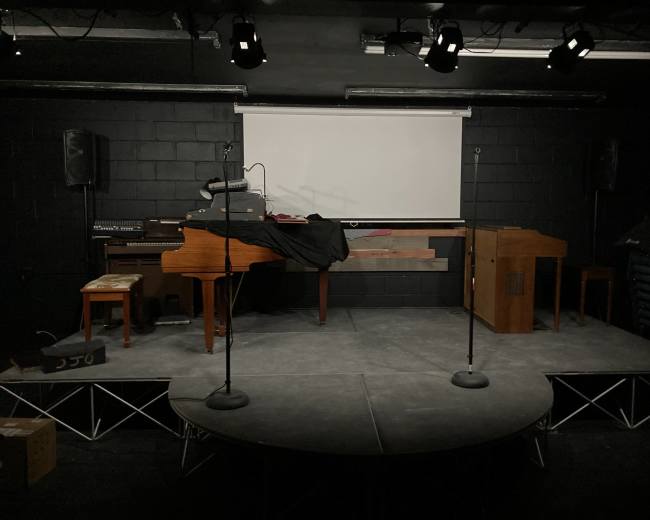 The image shows a stage setup with a grand piano, an organ, and microphones, against a black backdrop with a projection screen.