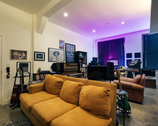 The image shows a cozy living room with a mustard-colored sofa, shelves filled with books and musical instruments, and a workspace featuring a piano and sound equipment under ambient lighting.