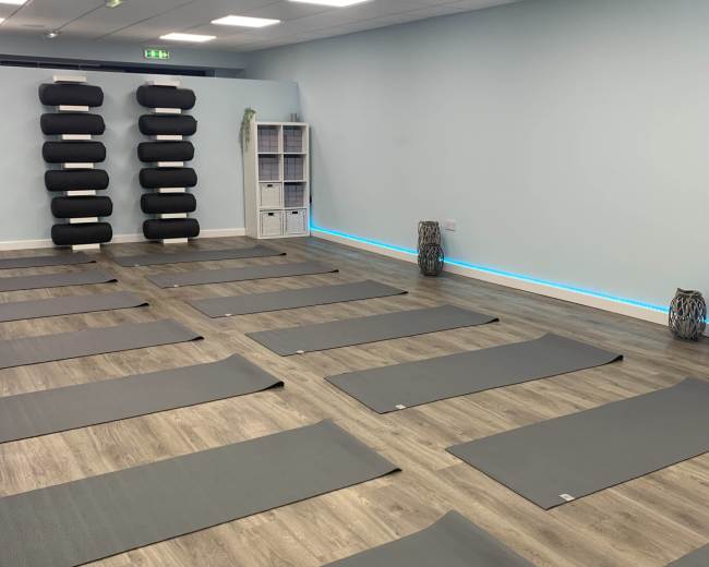 A spacious yoga studio with several gray mats arranged on the wooden floor and black yoga props stacked on a wall.