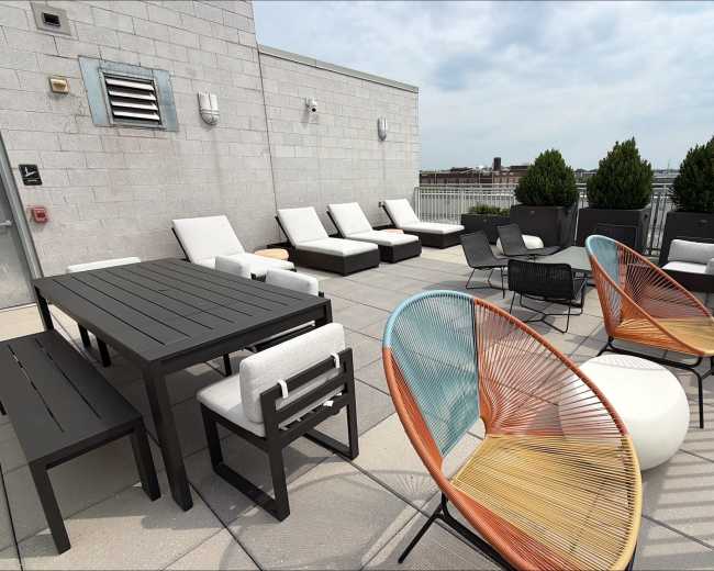 The rooftop terrace features modern lounge furniture, including a dining table, sun loungers, and colorful chairs, with potted plants lining the edges.