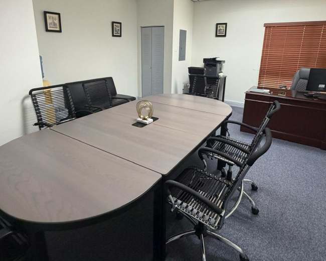 The image depicts a modern office space featuring a large conference table surrounded by black rolling chairs, with a desk and a window covered by blinds in the background.