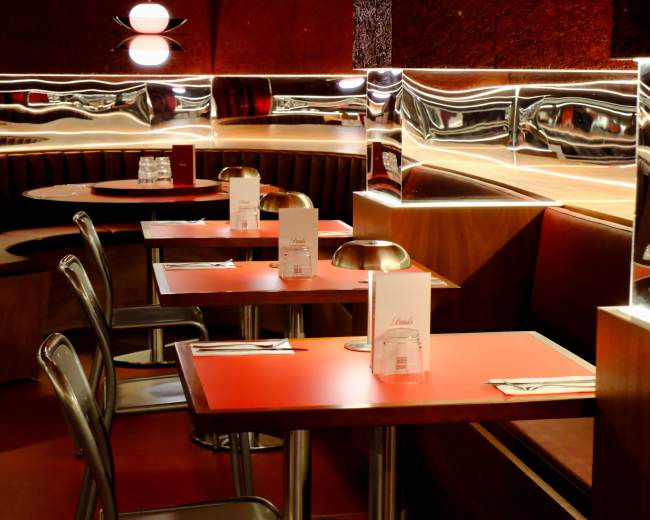 The image depicts a modern restaurant interior with sleek red tables, metallic accents, and curved seating areas.