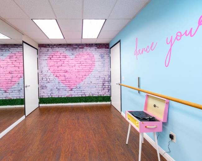 The image shows a dance studio with mirrored walls, a pink heart mural on a brick background, and a pink record player on a ballet bar.