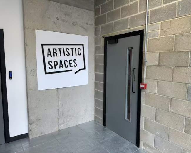 The image shows an industrial-style hallway with a large sign that reads "ARTISTIC SPACES" next to a gray door.