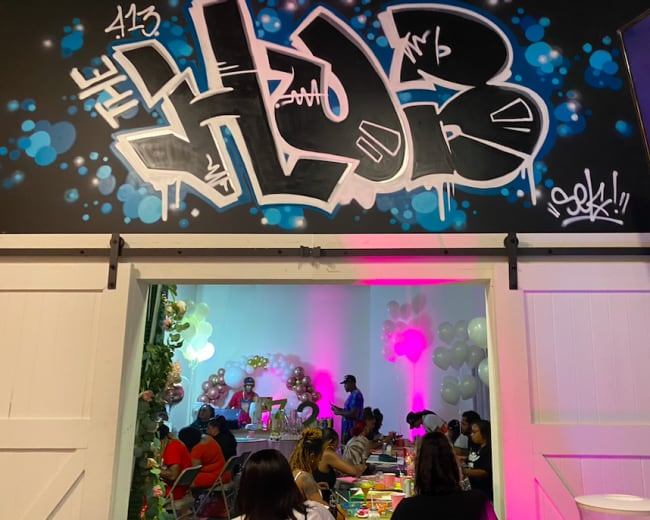 A colorful graffiti mural reading "THE HUB" adorns the wall above a gathering of people seated at tables decorated with balloons and party supplies.