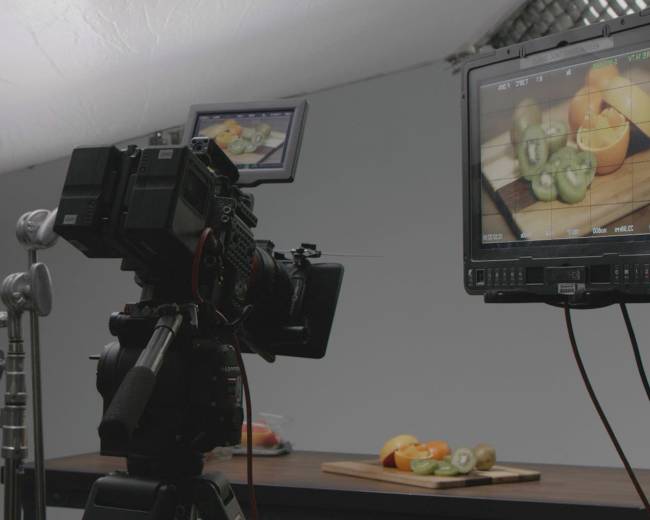 A camera is set up on a tripod, facing a monitor displaying a close-up of various fruits arranged on a wooden cutting board.