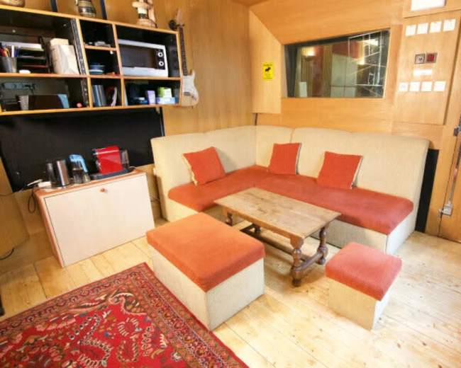 The image shows a cozy wooden room with a sofa, a coffee table, and a small area for appliances along with shelves and a carpet.