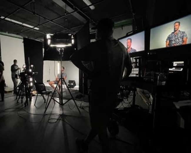 A film crew sets up equipment in a dimly lit studio with a subject seated under bright lights and two monitors displaying their image.