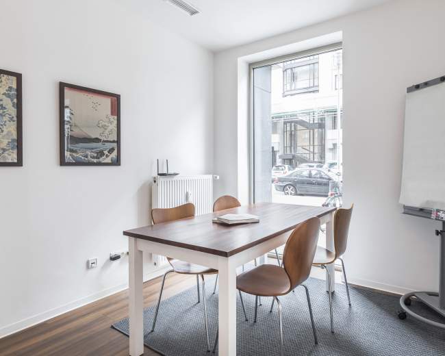 A small conference room features a wooden table with four chairs, a flip chart, and a window overlooking a street.
