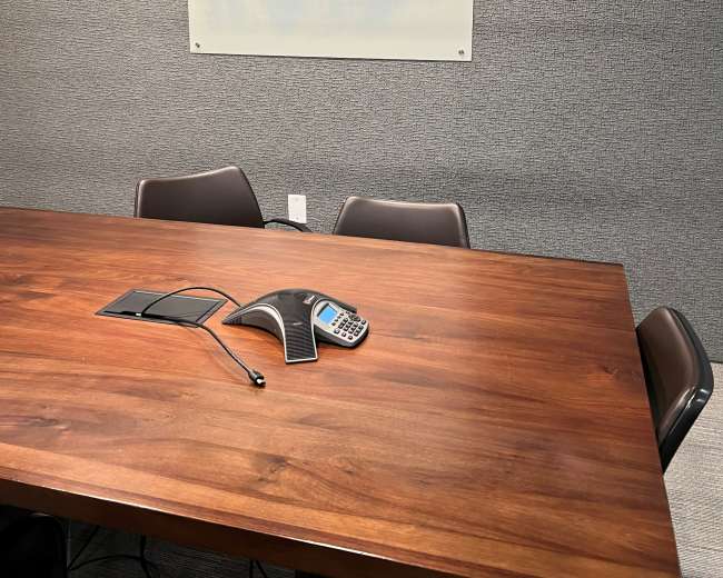 The image shows a conference room with a wooden table, a teleconferencing device, and a clear glass whiteboard mounted on the wall.