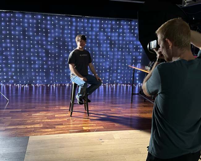 A person sits on a stool in front of a backdrop of string lights while another person films with a camera.