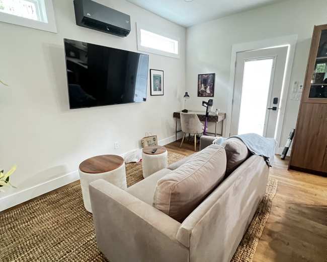 A living room features a sofa facing a mounted television, with a small table and workspace near a doorway.