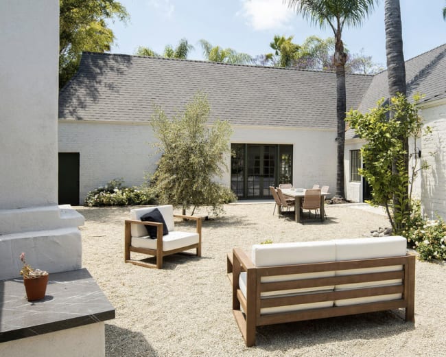 A minimalist outdoor patio features wooden furniture and a gravel surface, surrounded by greenery and a white brick building.