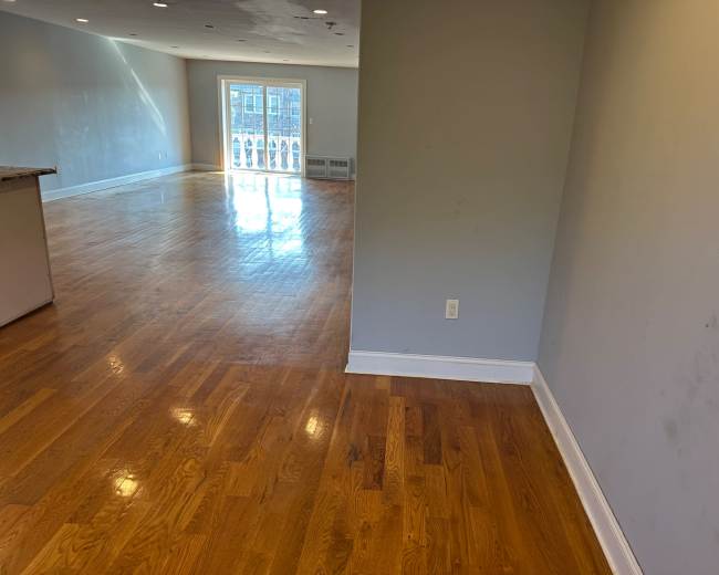 The image shows a spacious room with polished hardwood floors, leading towards a doorway that opens to a balcony.
