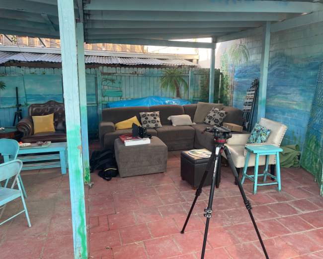The scene shows a partially sheltered outdoor space furnished with a gray sofa, chairs, and a tripod.