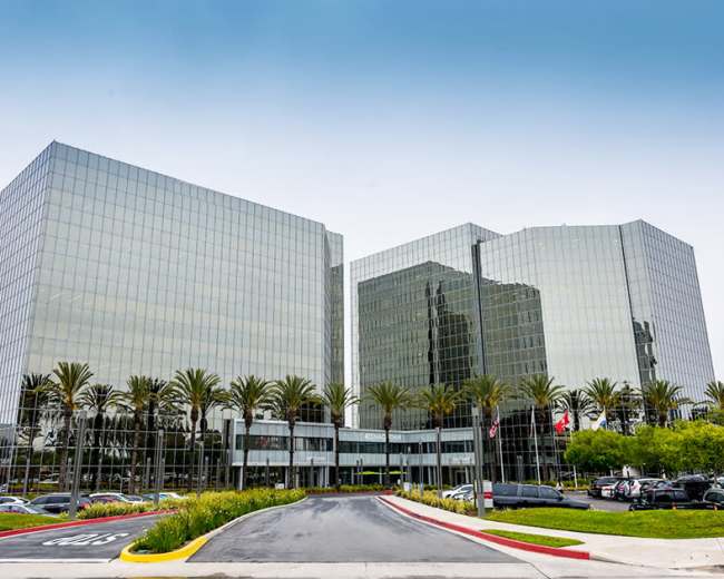 The image shows a modern glass office building with palm trees in the foreground and a parking area in front.