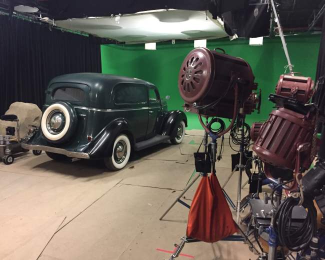 A vintage car is parked on a film set in front of a green screen, surrounded by lighting and camera equipment.