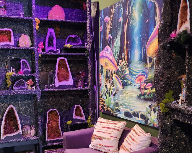 The image shows a cozy corner with a purple couch against a wall adorned with colorful crystal displays and a whimsical forest mural.