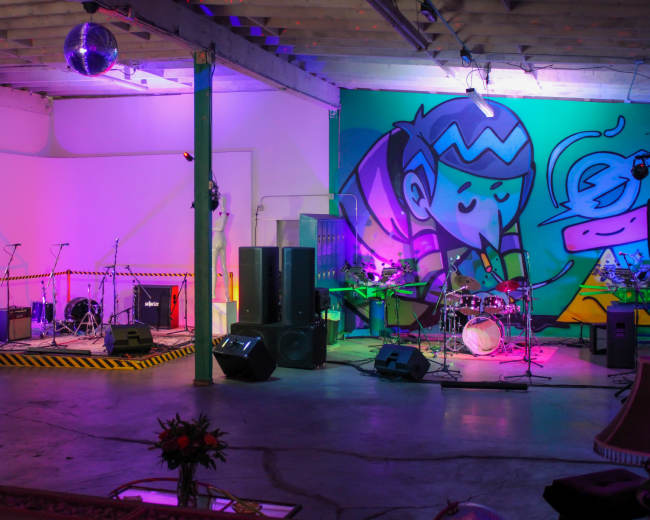 A colorful indoor venue set up for a performance, featuring a stage with musical instruments, amplifiers, and a large graffiti mural on the wall.