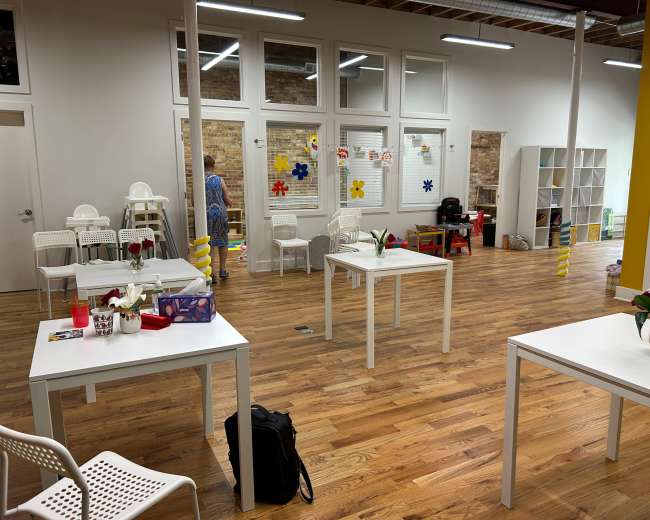 The image shows a bright, spacious room with wooden floors, several white tables, and chairs, along with colorful decorations on the walls and some children's toys.