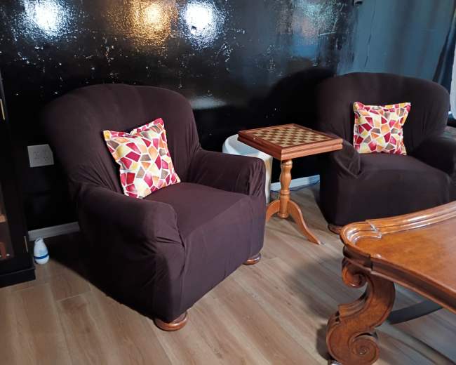 The image shows two dark, upholstered chairs with decorative pillows beside a small wooden side table featuring a checkerboard pattern in a room with a black wall.