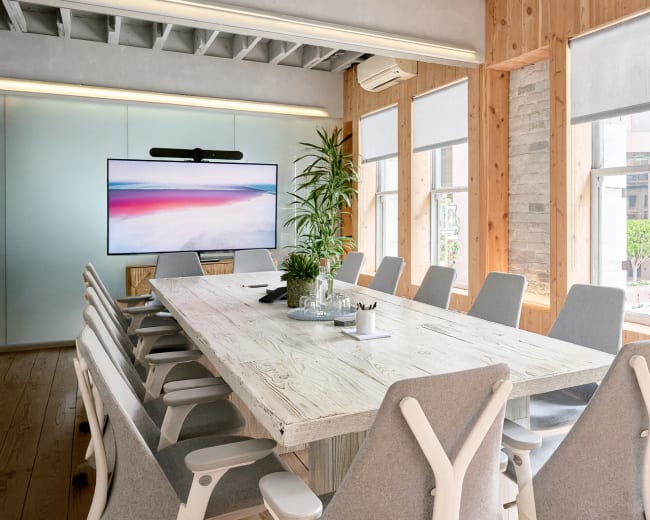 The image shows a modern conference room with a large wooden table, ergonomic chairs, and a screen displaying a landscape, illuminated by natural light through large windows.