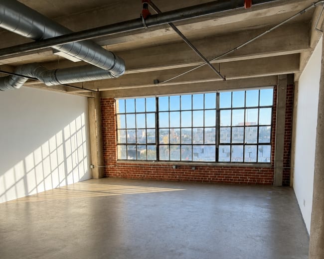 The image shows an industrial-style room with large windows allowing natural light to enter, exposing brick walls and concrete flooring.