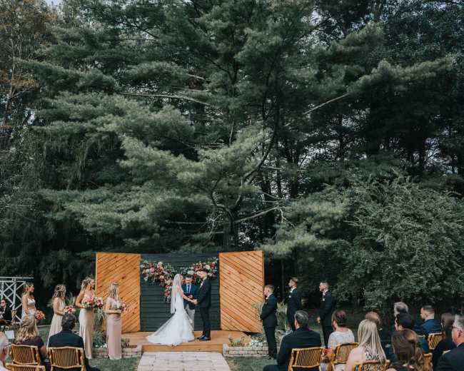 A wedding ceremony takes place outdoors on a wooden platform, surrounded by guests seated in rows and flanked by trees.