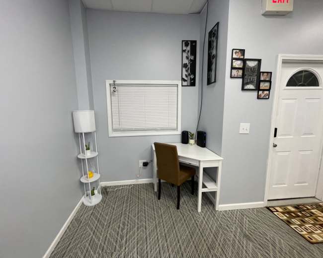 The image shows a small room with a desk and a chair beside a window, a lamp on a stand, and a door leading outside.