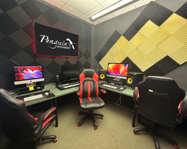 A music production studio featuring three workstations with computers, keyboards, and studio monitors, surrounded by sound-absorbing wall panels.