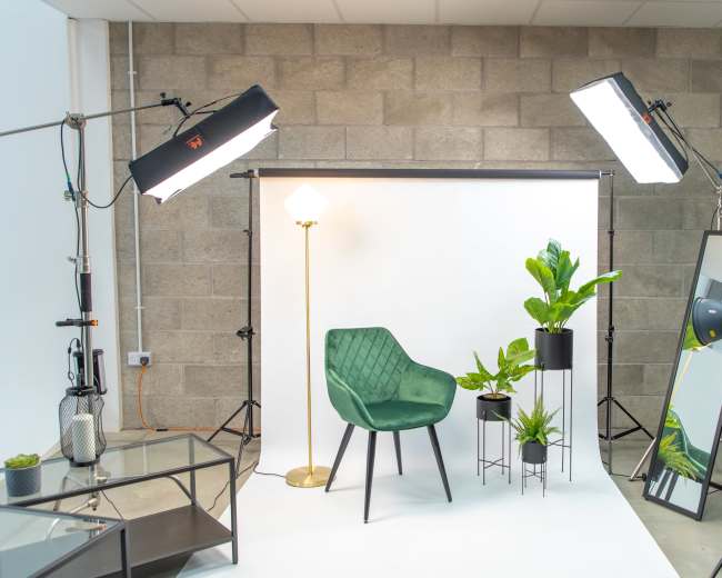 A photo studio setup featuring a green chair, a floor lamp, two potted plants, and various lighting equipment against a backdrop of white and gray walls.