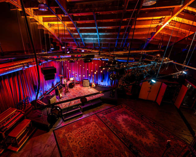 The image shows a dimly lit performance space with vibrant colored lights, a raised stage, and musical equipment arranged on the floor.
