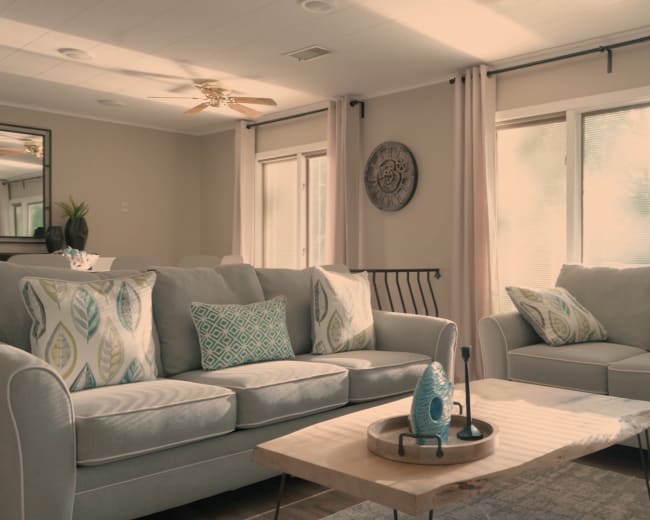 A light-filled living room features two gray sofas adorned with decorative pillows, a wooden coffee table, and large windows dressed with sheer curtains.
