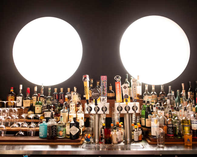 A bar counter is lined with various bottles of alcohol, with two large circular lights illuminating the scene.