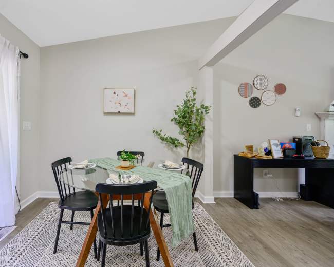 A dining area features a round table set for four, surrounded by black chairs, with a green table runner, plants, and decorative wall art.
