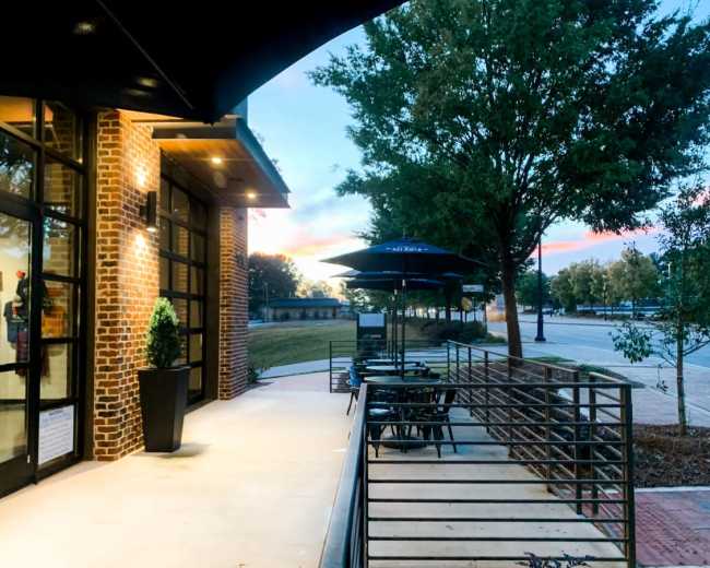 The image shows an outdoor seating area with metal tables and umbrellas beside a brick building, framed by trees and a colorful sky at dusk.