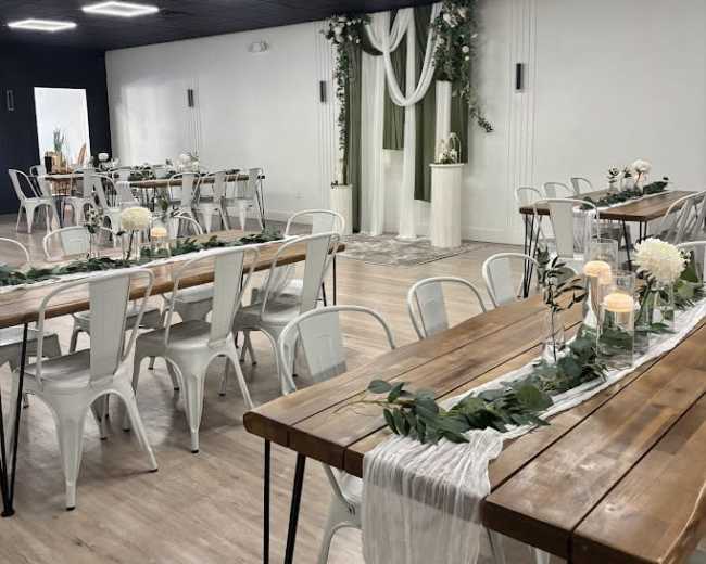 The image shows an event space arranged with wooden tables and white chairs, decorated with greenery and floral centerpieces.