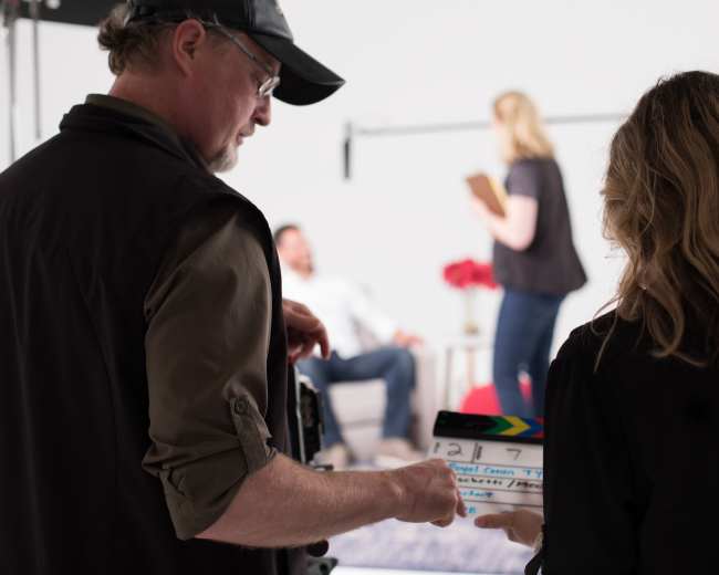 A man and a woman prepare to film a scene with a clapboard in a studio setting, while two actors are visible in the background.