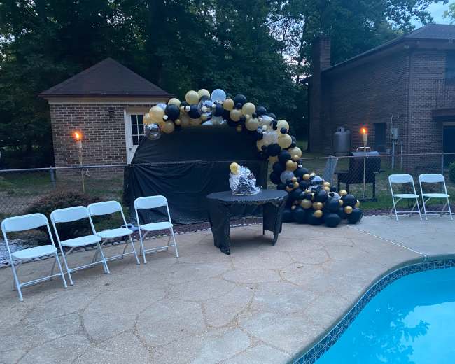 A decorated seating area with a table is set up by a swimming pool, featuring a balloon arch in black and gold colors nearby.