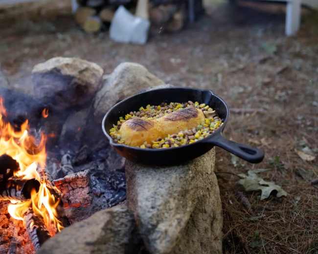 A cast-iron skillet with food cooks over a campfire, surrounded by a stone fire pit and wooden chairs, with a red barn and stacked firewood in the background.