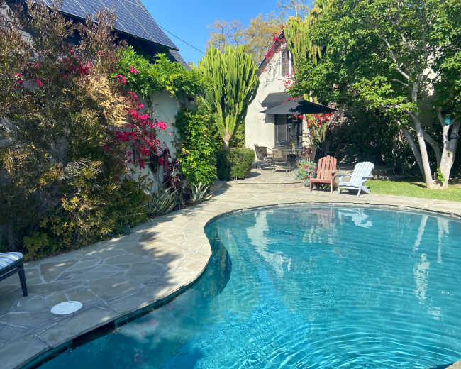 A landscaped backyard with a swimming pool surrounded by stone paving and colorful plants, featuring two lounge chairs and a patio area in the background.