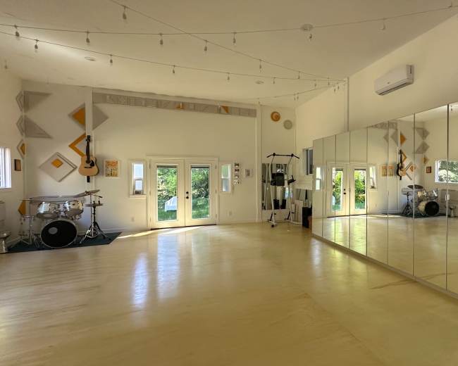 The image shows a spacious dance or practice studio with large mirrors, wooden floors, and natural light coming through multiple windows.
