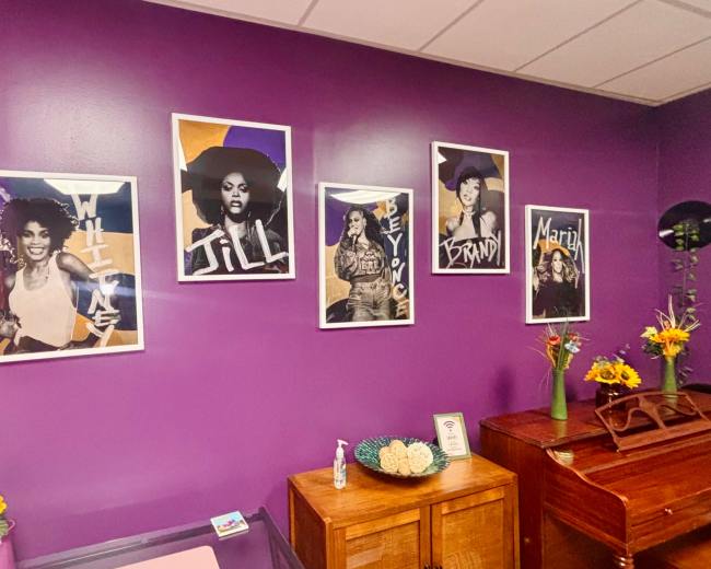 The image shows a purple wall adorned with five framed portraits of women, accompanied by flowers on a wooden table in front.