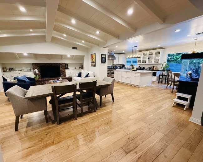 The image shows a spacious, inviting living area with a dining table, comfortable seating, and an open kitchen, featuring wooden floors and exposed beam ceilings.