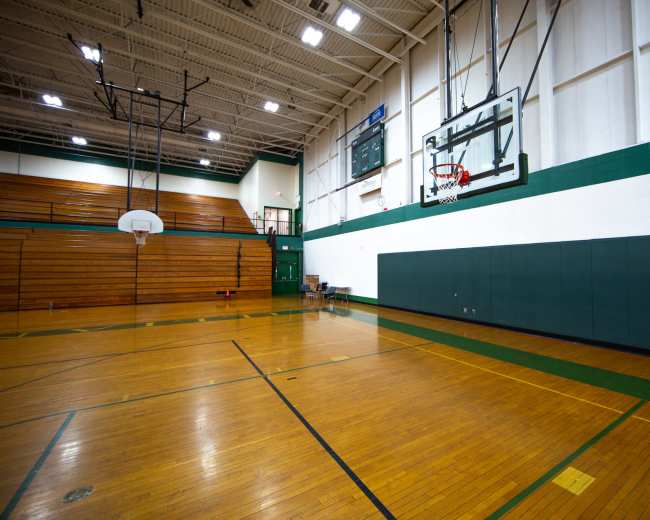 The image shows an empty gymnasium with a basketball hoop on one side and wooden bleachers along the back wall.