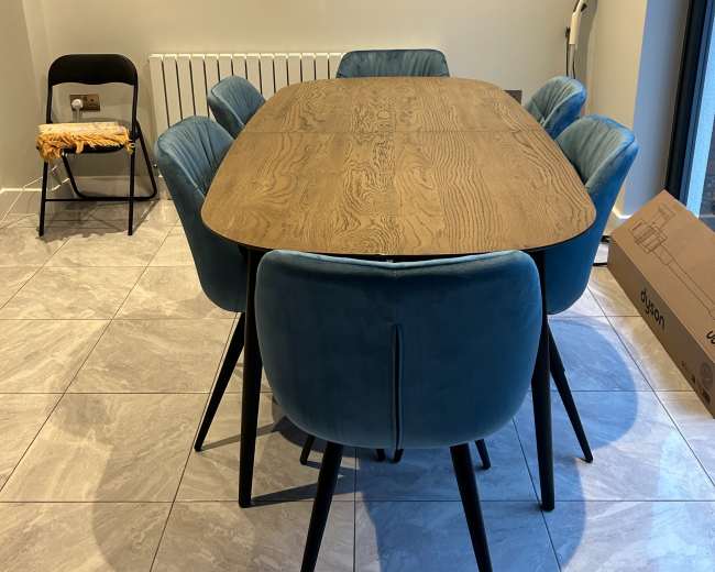 A wooden dining table with blue upholstered chairs is set in a room with tiled flooring and a wall-mounted heater.