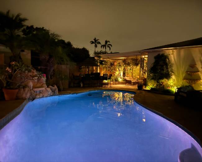A illuminated swimming pool with surrounding palm trees and a patio area featuring seating and plants under a night sky.