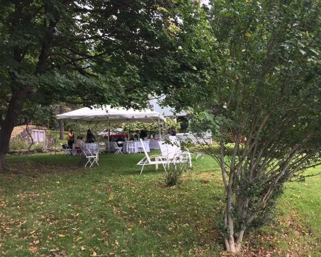 A large white tent is set up in a grassy area, surrounded by trees and chairs arranged for an outdoor gathering.