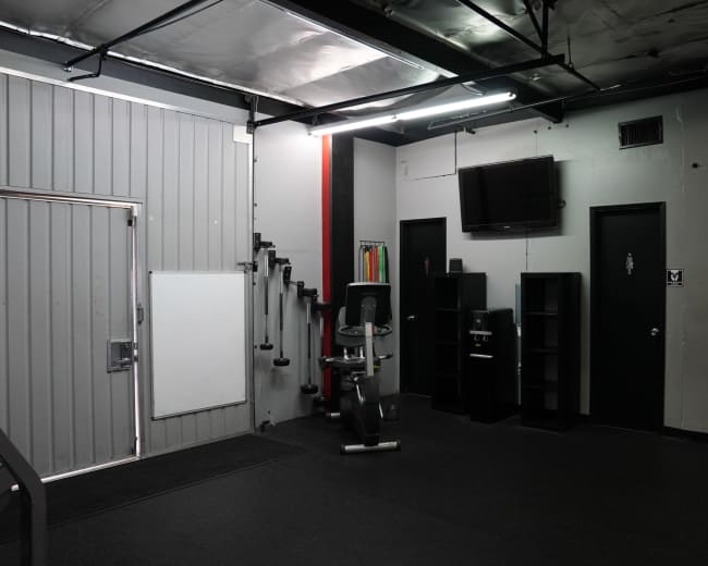 The image shows a corner of a gym featuring exercise equipment, a wall-mounted television, and storage shelves against a gray wall with a black door.