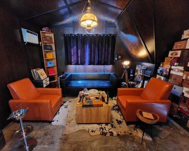 The image shows a cozy room with two orange chairs facing a wooden coffee table, surrounded by shelves and boxes, and illuminated by a pendant light.